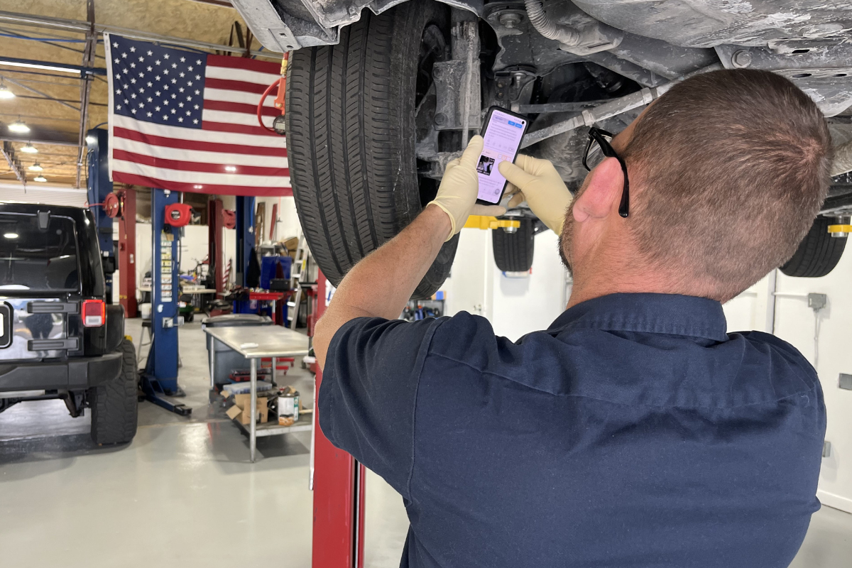Technician performing a Digital Vehicle Inspection (DVI) on a car in Boca Raton, FL. The image shows the technician under the car with a phone, capturing detailed photos and videos of the vehicle's condition for a comprehensive and transparent inspection report at Boca Auto Fix.