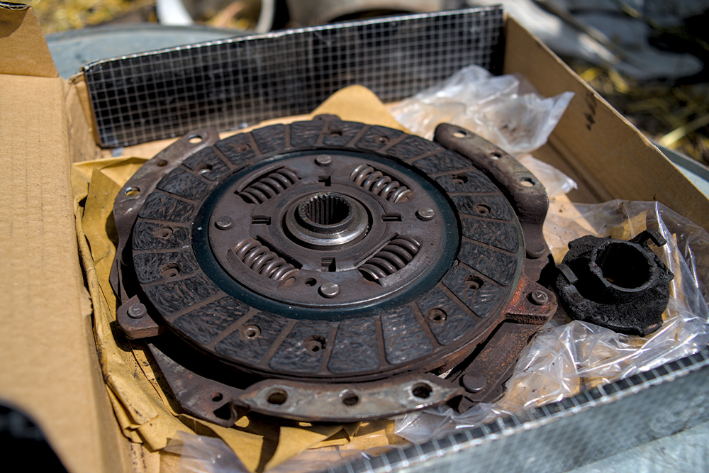A close-up of a rusty automotive clutch disc in a cardboard box with packaging material and a worn rubber component nearby.