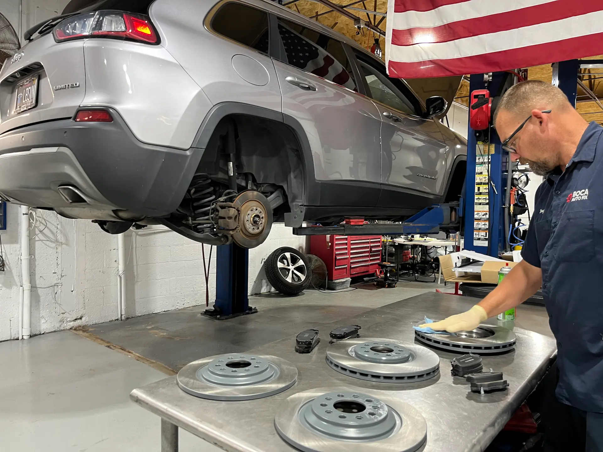Mechanic in an auto shop cleans a brake rotor from a raised Jeep with wheels removed.