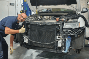 Cooling system repair in Boca Raton, FL, at Boca Auto Fix. Image of Doug DeLucca stands to the left of a white SUV with its front bumper removed. The vehicle's hood is propped open, exposing the engine bay, radiator core, cooling system, and internal structural components in an automotive repair shop setting.