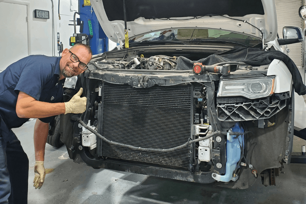 Cooling system repair in Boca Raton, FL, at Boca Auto Fix. Image of Doug DeLucca stands to the left of a white SUV with its front bumper removed. The vehicle's hood is propped open, exposing the engine bay, radiator core, cooling system, and internal structural components in an automotive repair shop setting.